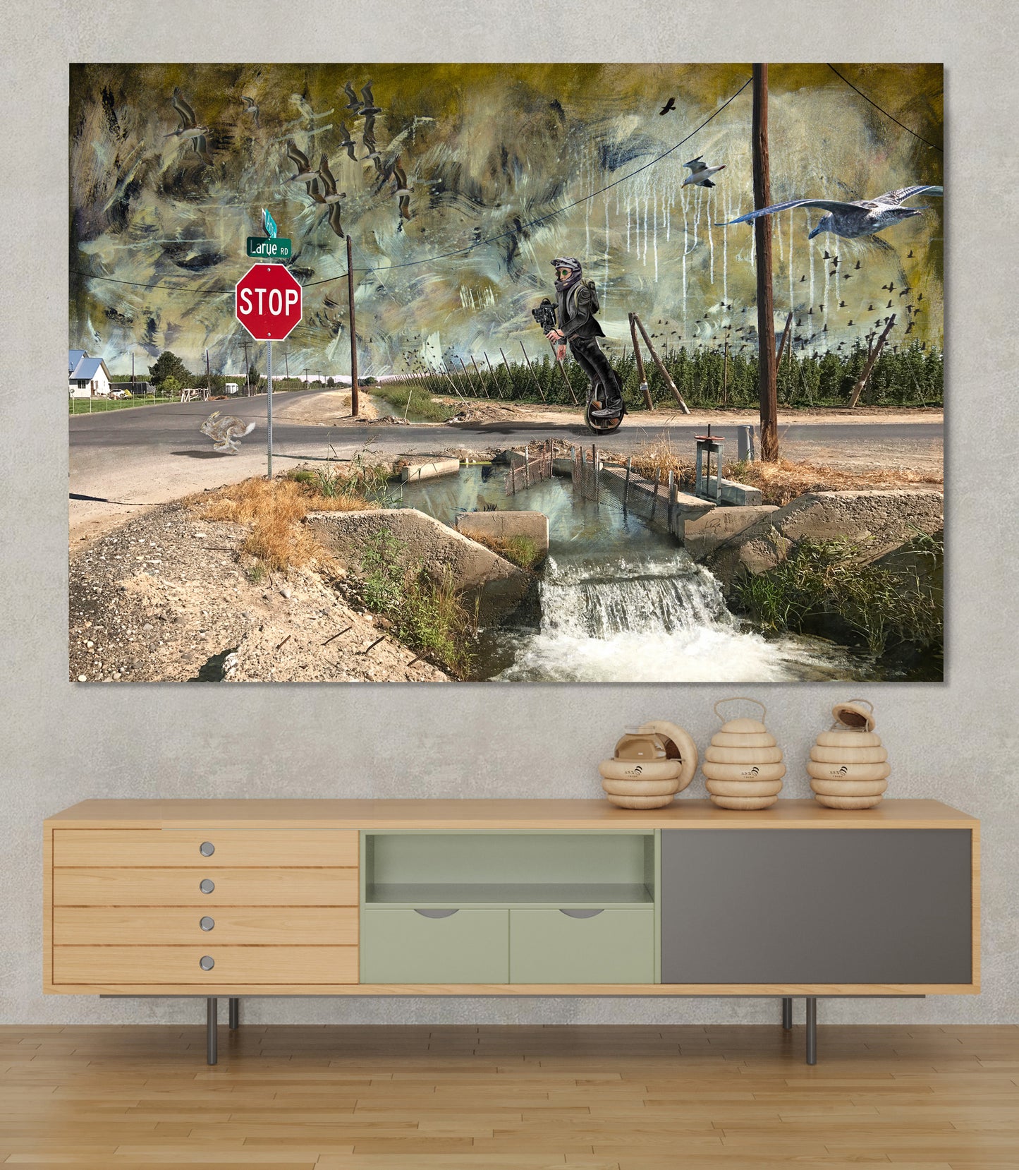 A large rectangular wall art print depicting an abstract outdoor scene with a stop sign, water channel, birds, and a dark central figure, displayed above a light wood console cabinet in a styled interior. LaRue Road bye Doug LaRue
