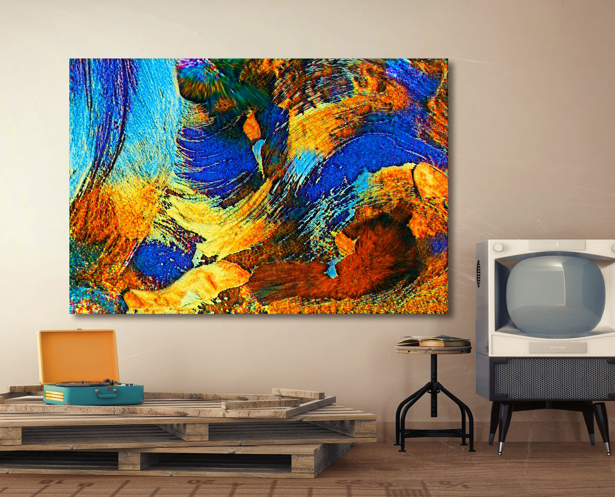 A large square abstract canvas print with vibrant blue, orange, yellow, and brown brushstrokes, displayed on a beige wall in a styled room setting with a wooden pallet coffee table, a vintage television, and a small stool. Rollingwood Cobalt by Doug LaRue