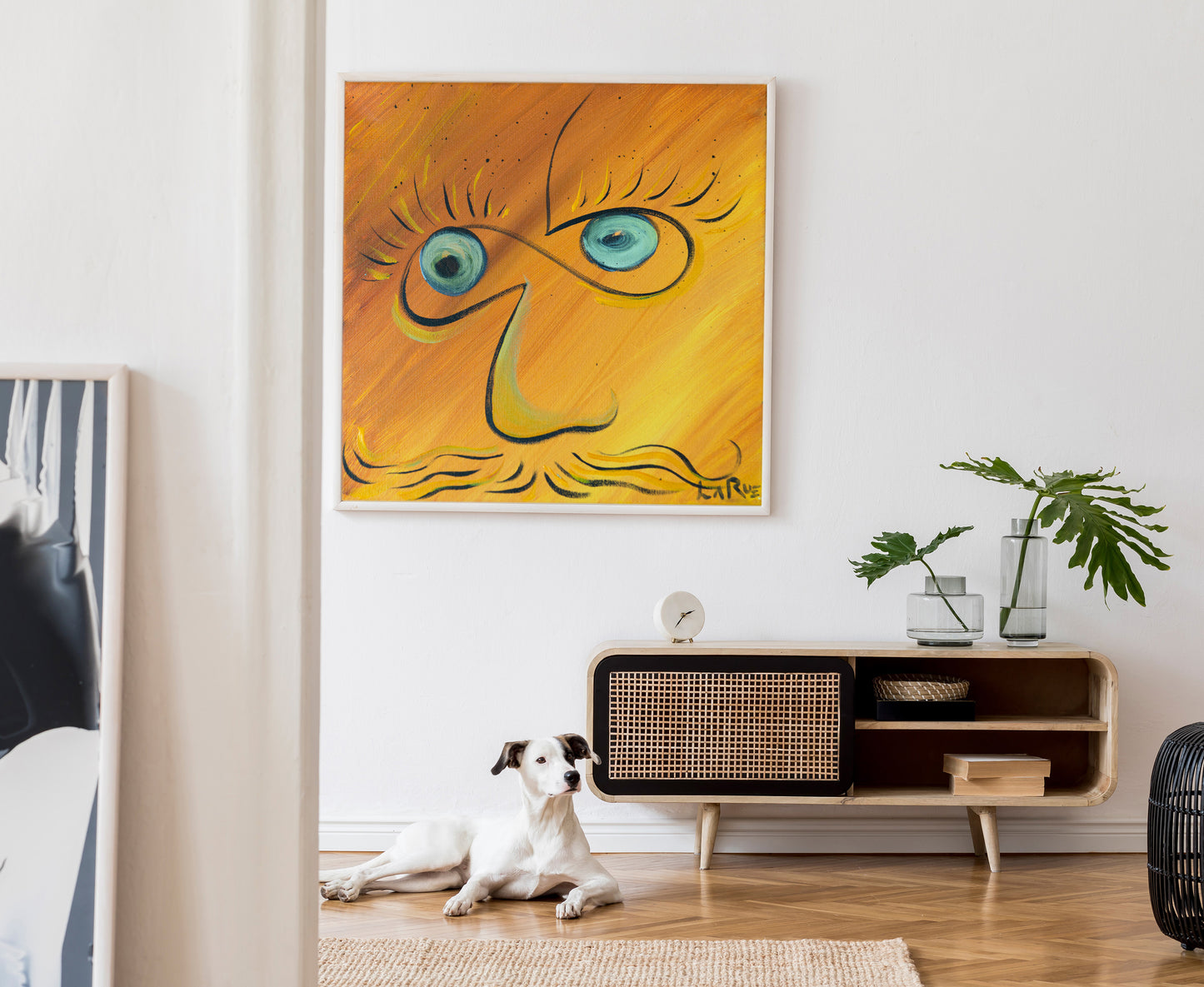 A framed abstract painting with a stylized face in yellow, orange, and blue hangs on a white wall above a wooden sideboard. A white dog lies on a woven rug on the floor below, and a potted plant sits on the cabinet. Infinite Eyes #1 by Doug LaRue