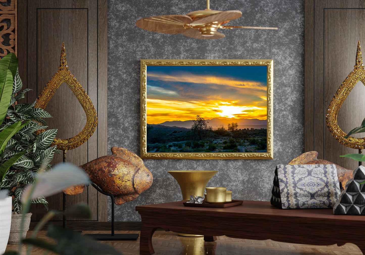 A framed landscape painting of a desert sunset is displayed on a textured gray wall in a styled room setting with a wooden table, decorative pillows, plants, and gold-toned wall mirrors.