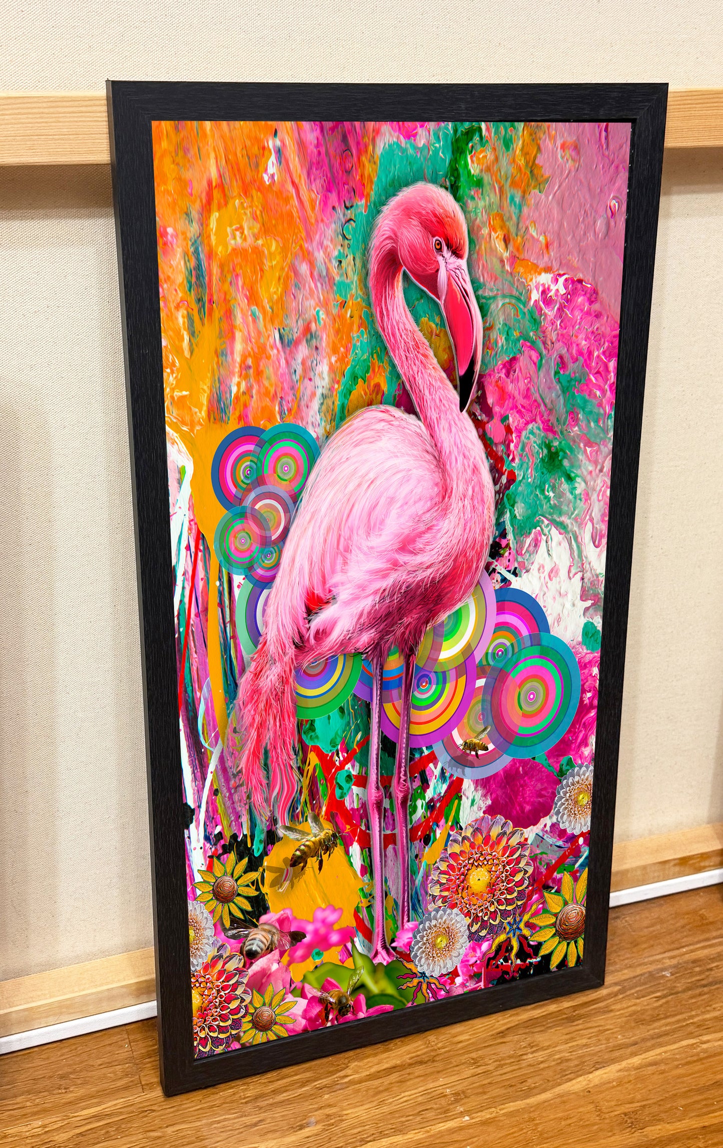 A framed vertical art print featuring a bright pink flamingo surrounded by colorful abstract floral and circular patterns, displayed leaning against a beige wall on a wooden floor.