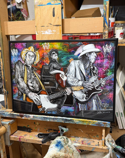 A framed colorful art print depicting three guitarists in black and white against a vibrant multicolor painted background, displayed on an artist's workbench in a studio. Slim, Red and Stevie Ray by Doug LaRue
