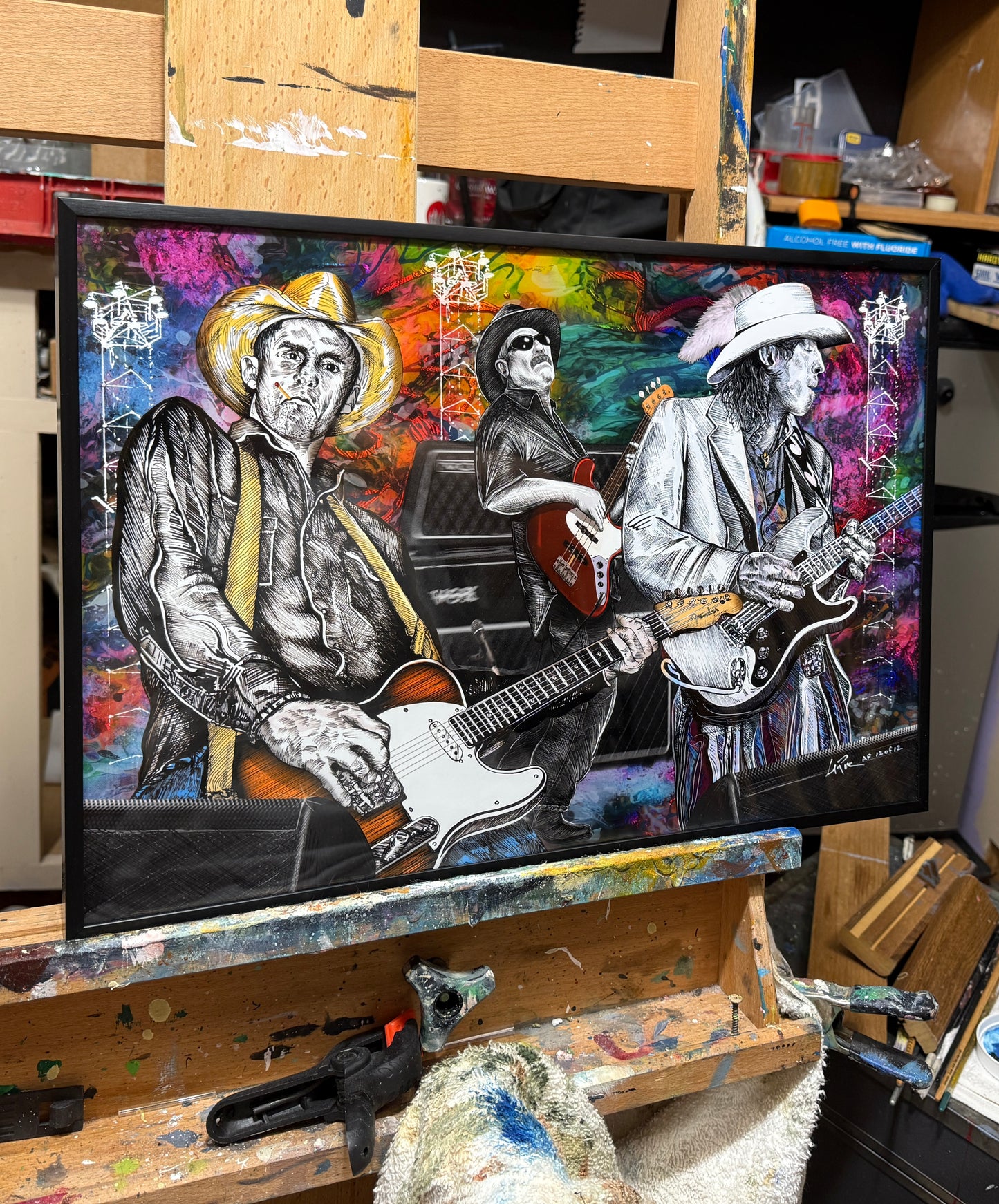 A framed colorful portrait painting of three guitarists on an easel in an art studio. The artwork features black-and-white illustrated figures with vivid multicolor painted backgrounds in pink, blue, purple, orange, and green. Slim, Red and Stevie Ray by Doug LaRue