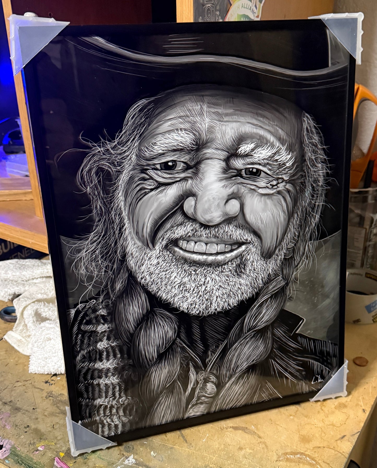 Black and white portrait of a smiling Willie Nelson portrait by Doug LaRue on a dark background, displayed on a wooden surface.
