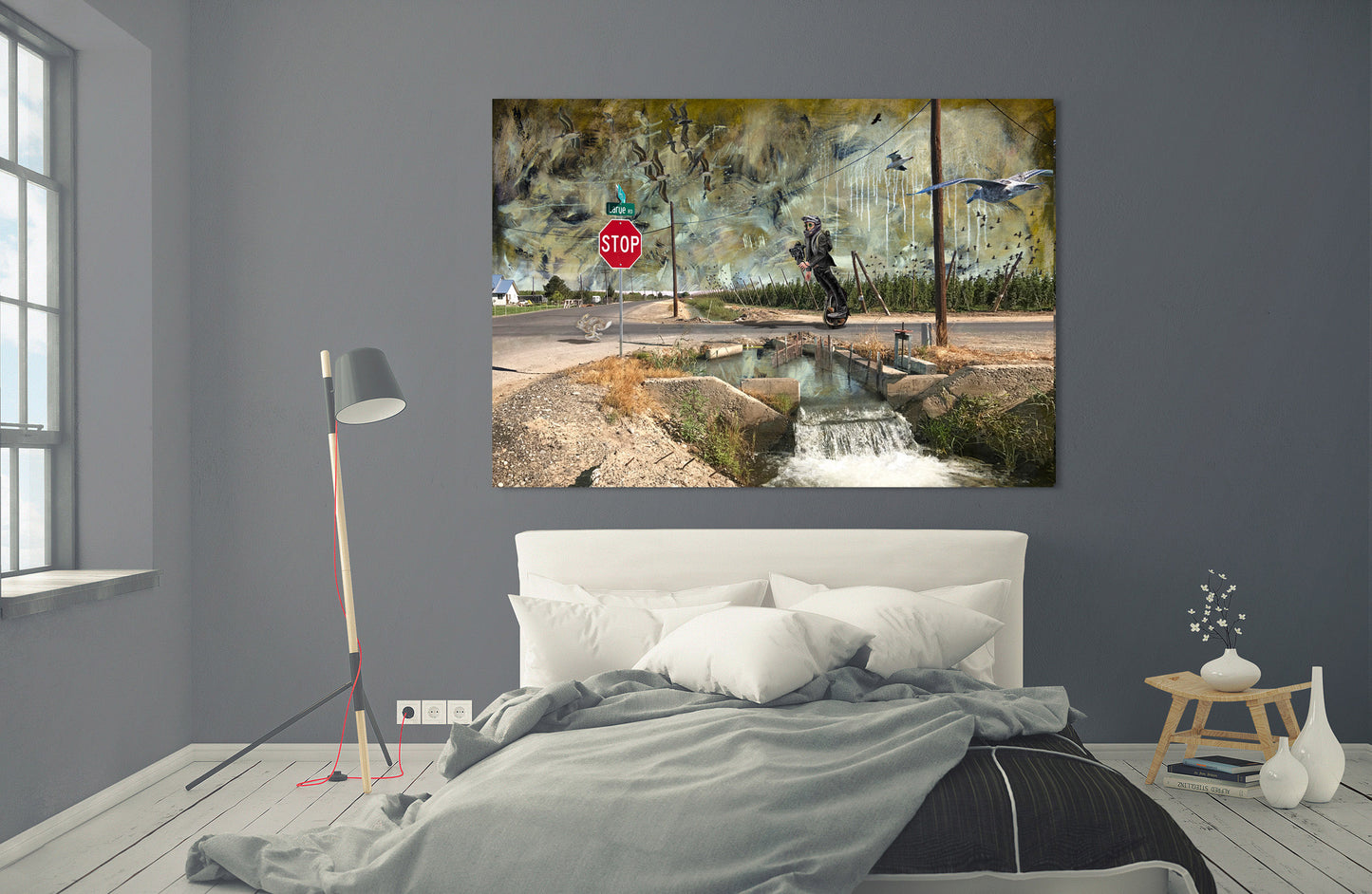 A canvas wall art print depicting a rustic watermill and stream scene with a stop sign, displayed on a dark gray wall above a neatly made bed in a modern bedroom setting. LaRue Road bye Doug LaRue