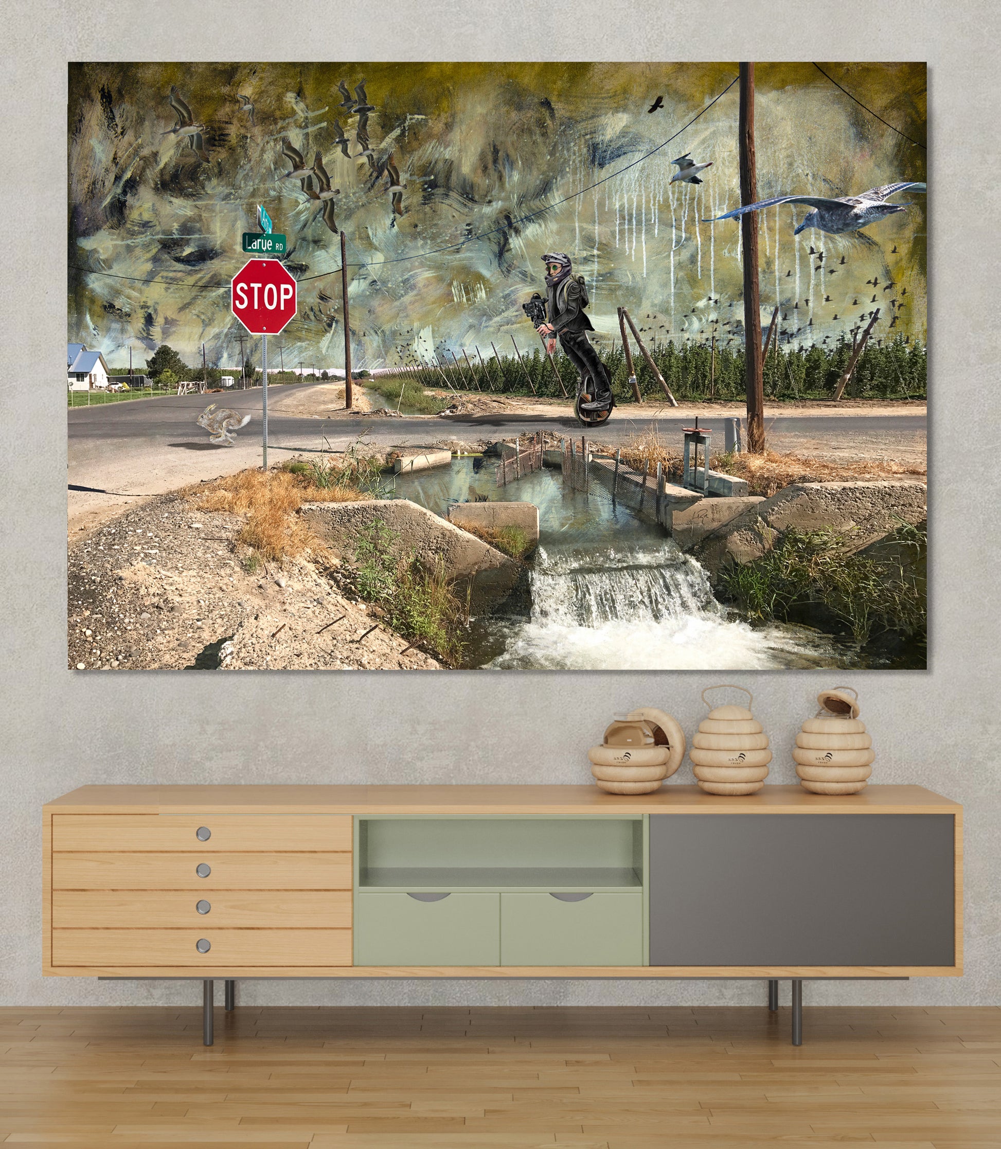 A large rectangular wall art print depicting an abstract outdoor scene with a stop sign, water channel, birds, and a dark central figure, displayed above a light wood console cabinet in a styled interior. LaRue Road bye Doug LaRue