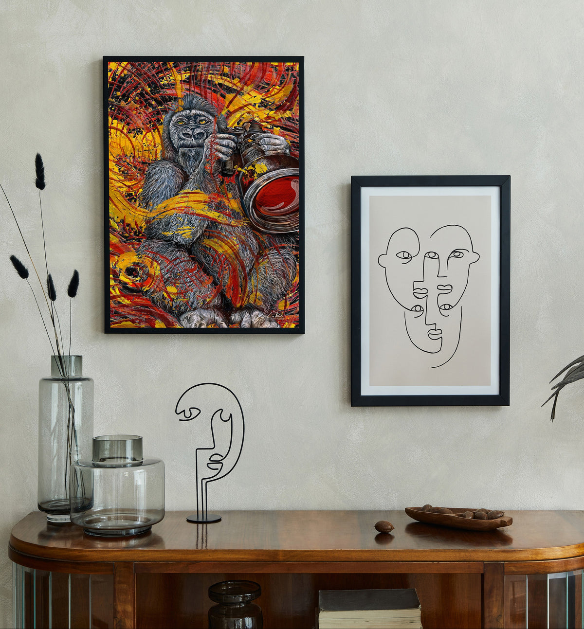 A styled interior scene showing two framed wall art pieces hanging above a wooden console table. The left frame contains a vivid abstract painting with a gorilla and warm red, orange, yellow, and black tones. Nature Photographer by Doug LaRue