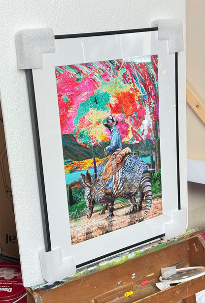 Colorful artwork of a cowboy riding a giant armadillo with a vibrant abstract sunset background. Armadillo Cowboy Sunset by Doug LaRue in a black metal frame with a white mat.
