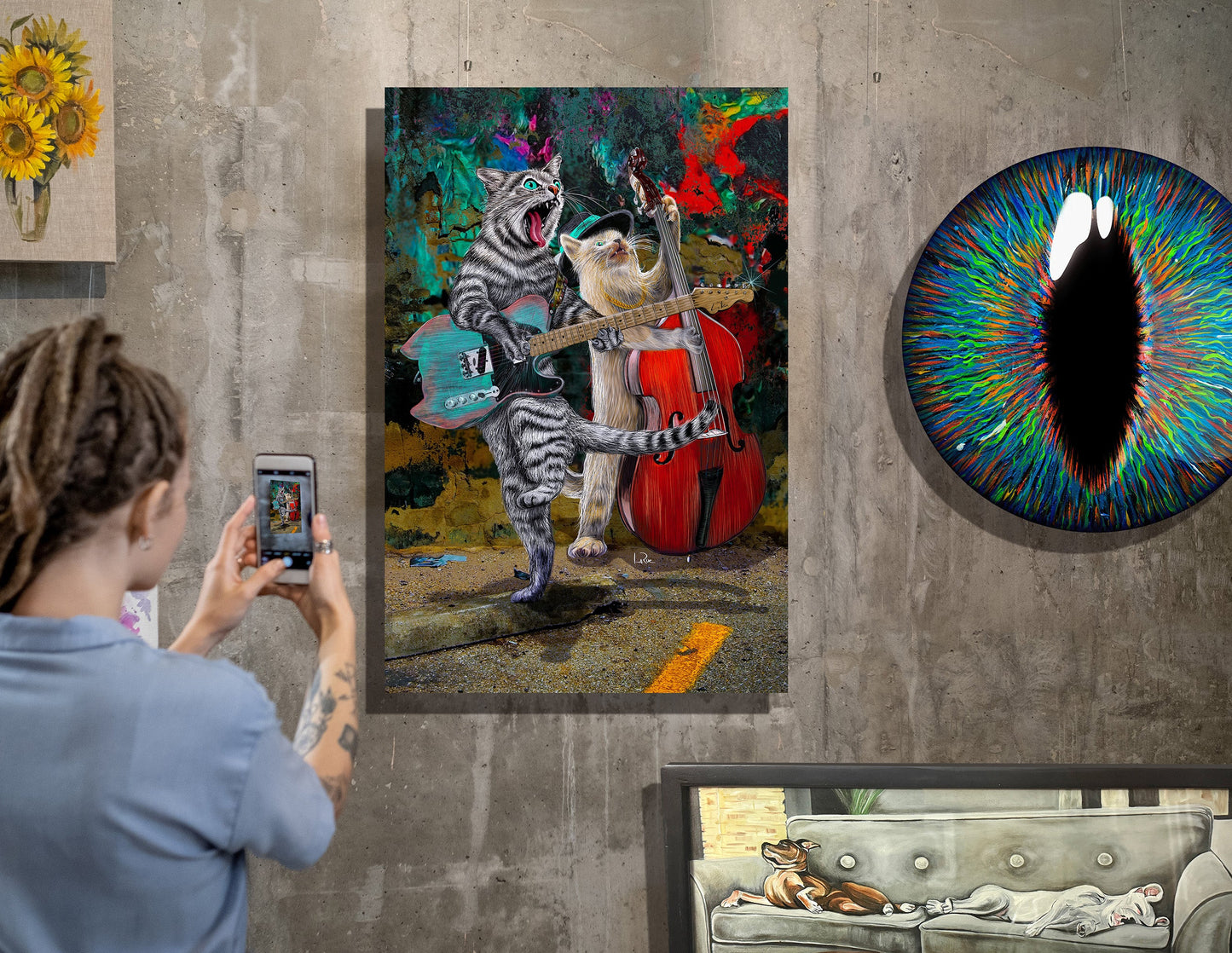 A person takes a photo of a colorful canvas print hanging on a gray concrete wall. The artwork depicts two cats playing string instruments together in a vibrant, multicolored scene.