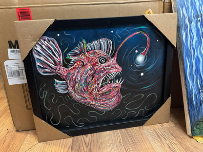 A canvas print featuring an original deep sea fish illustration, framed in a black museum style frame, displayed against a backdrop of packing materials.