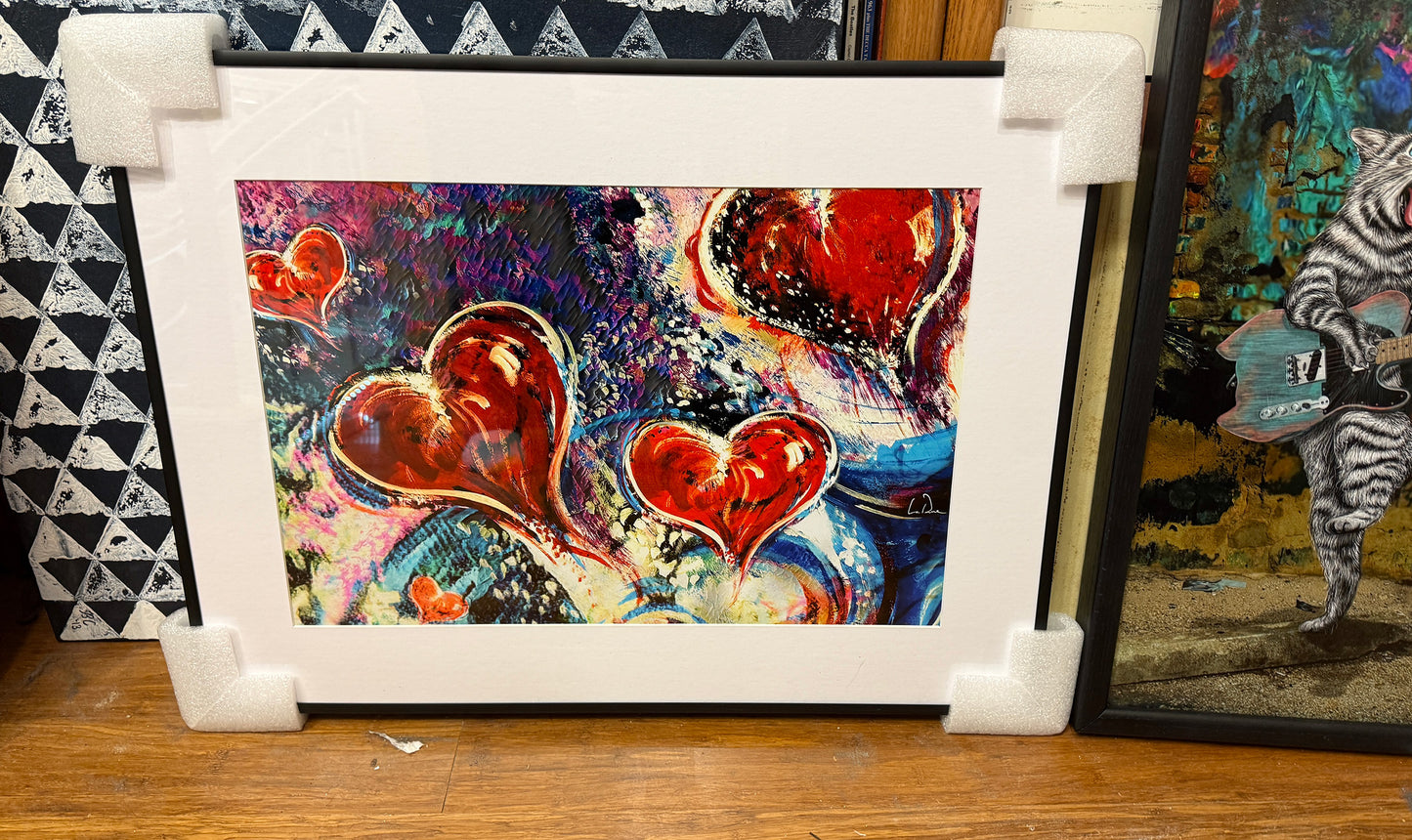 Colorful heart-themed artwork called Hearts Adrift by Doug LaRue in a white mat and black metal frame on a wooden surface with other artworks in the background.