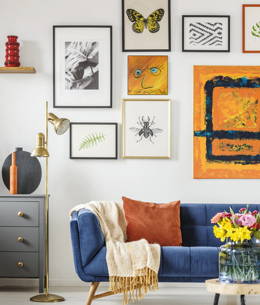 A styled living room corner featuring a gallery wall with multiple framed art prints above a blue velvet sofa. The wall displays a variety of colorful and monochrome artworks, including a butterfly print, a green fern leaf, a beetle illustration, and a large orange abstract painting. Infinite Eyes #1 by Doug LaRue
