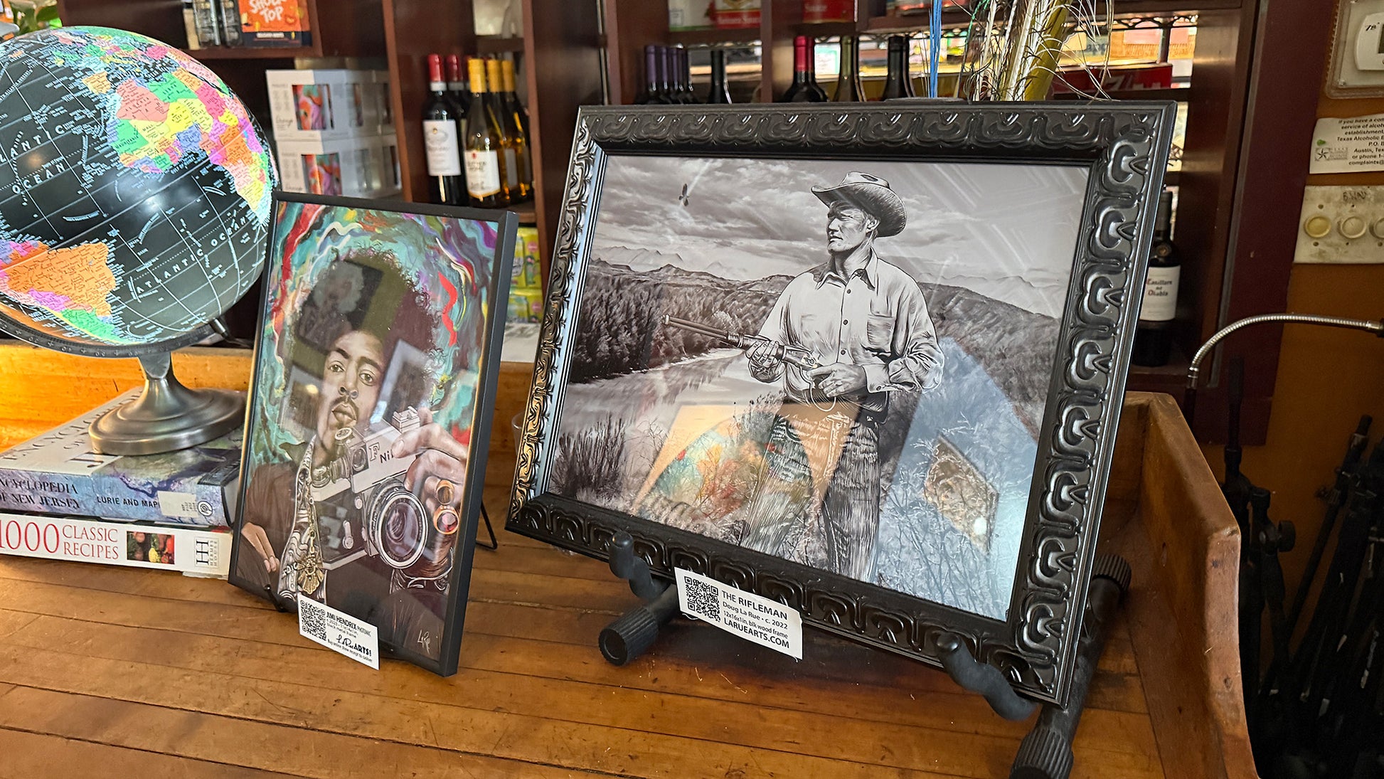 Framed artwork of a person with a hat on a wooden surface, surrounded by books and a globe. The Rifleman by Doug LaRue on the NeWorDeli Bar