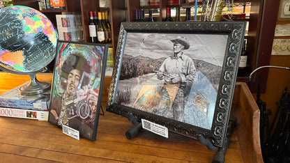 Framed artwork of a person with a hat on a wooden surface, surrounded by books and a globe. The Rifleman by Doug LaRue on the NeWorDeli Bar