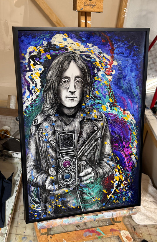 A framed portrait painting of John Lennon, depicted holding a camera, set against a vibrant abstract background with splashes of blue, purple, yellow, and white. The artwork is housed in a black frame and displayed on an easel in a studio or workshop environment.