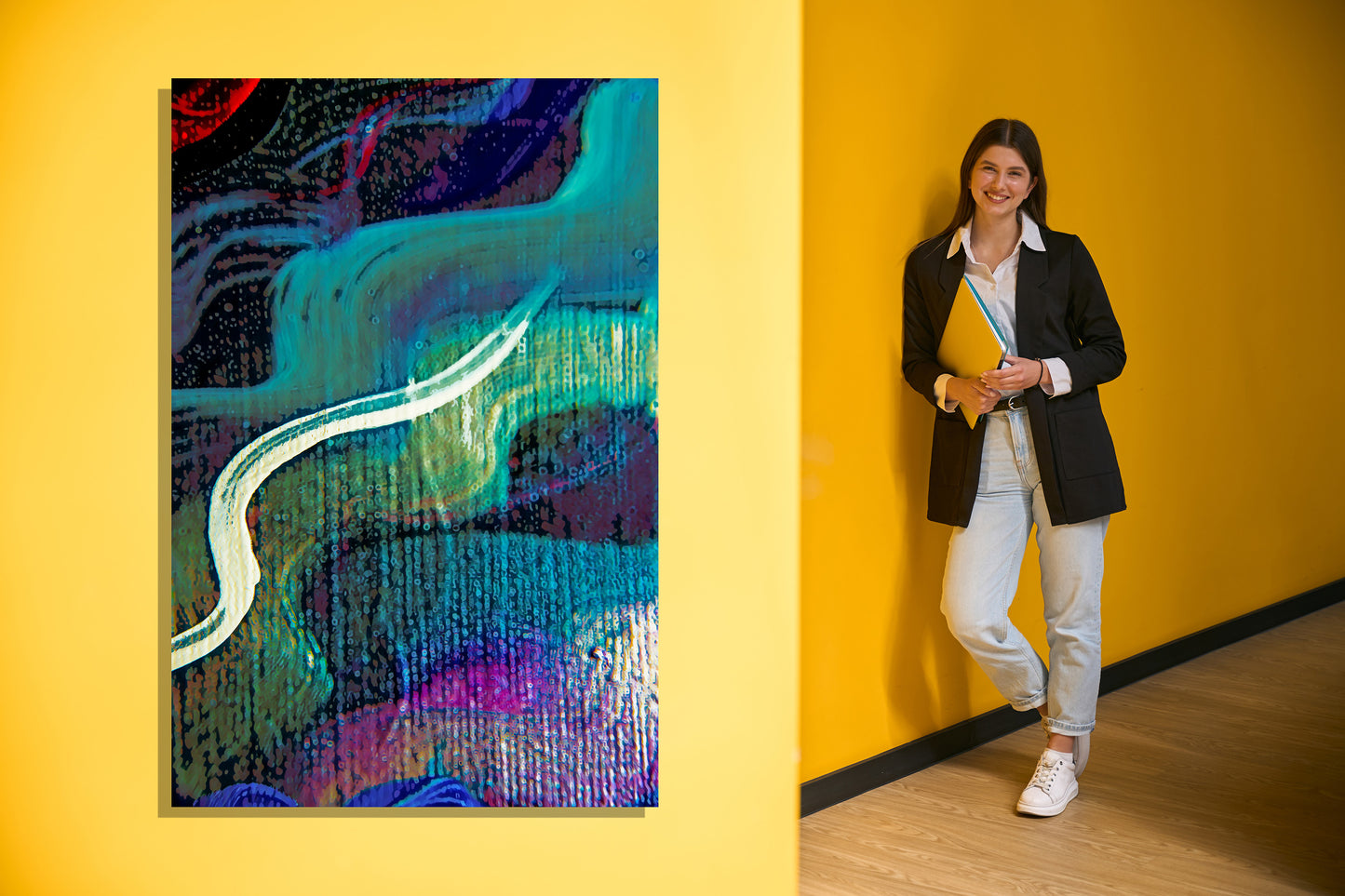 A woman in a black blazer, white shirt, and light jeans stands against a bright yellow wall holding a yellow folder, next to a colorful abstract painting on the same yellow wall.  Lost Creek art by Doug LaRue