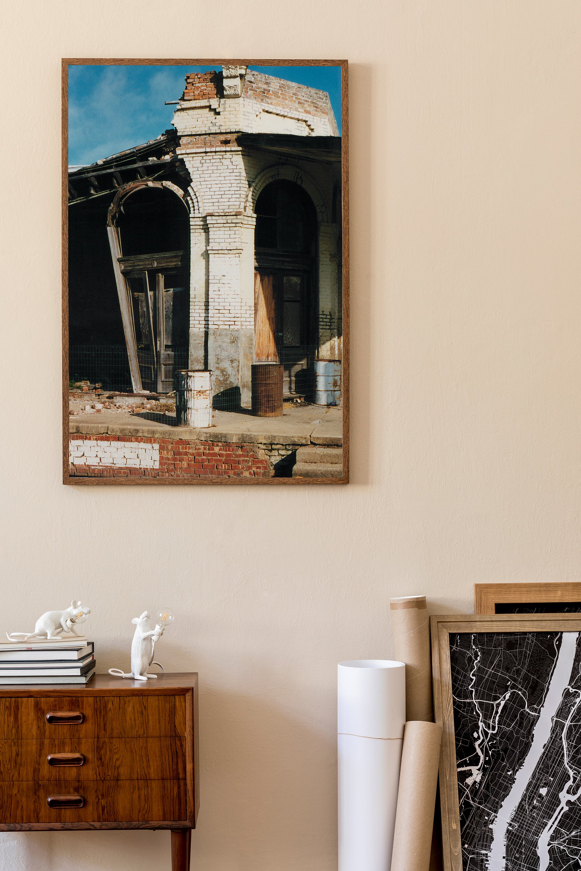 A framed art print depicting a weathered brick building with arches, hanging on a beige wall above a wooden cabinet in a styled interior setting.