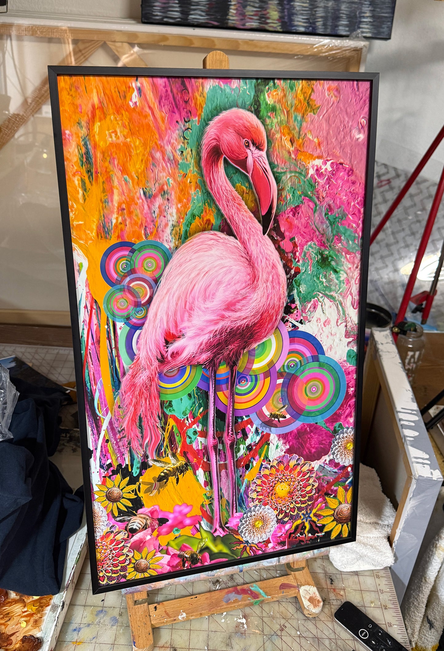 A framed colorful painting of a pink flamingo standing among bright floral and abstract patterns, displayed on a small easel in a workshop or studio setting. Pink Flamingo by Doug LaRue