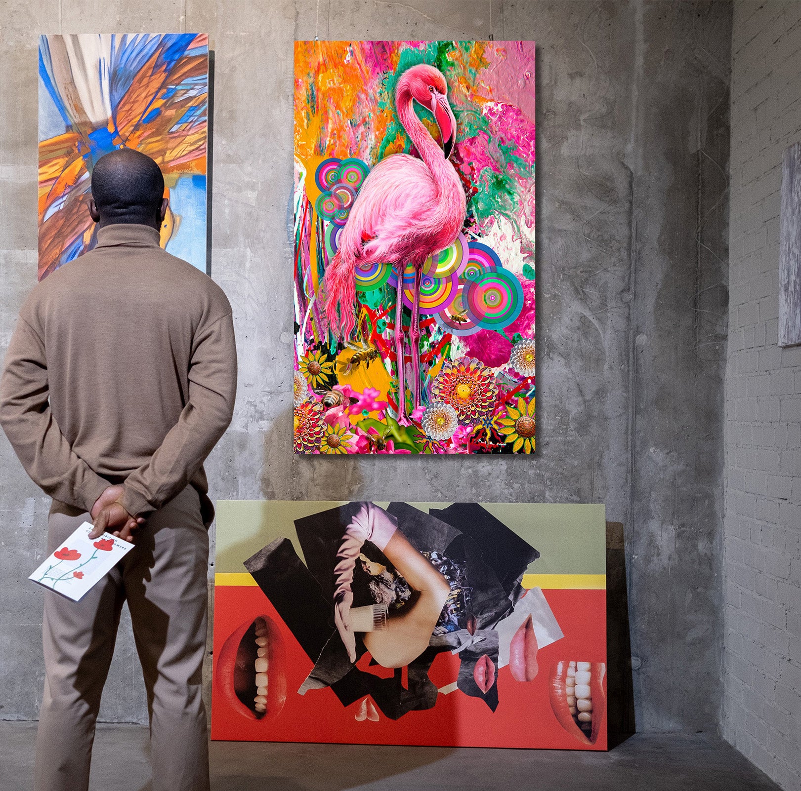A person stands with their back to the camera, viewing a large, highly colorful abstract painting of a pink flamingo on a gallery wall. Another colorful painting is visible on the left, and a large red and black artwork is leaning on the floor in the foreground. Pink Flamingo by Doug LaRue