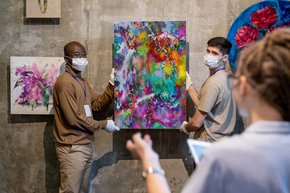 Two people wearing face masks hold up a large, vibrant abstract painting with a colorful mix of green, blue, red, yellow, and purple. Another person is visible in the foreground, and other paintings hang on a textured gray wall in the background. Balcones Pop Tart by Doug LaRue