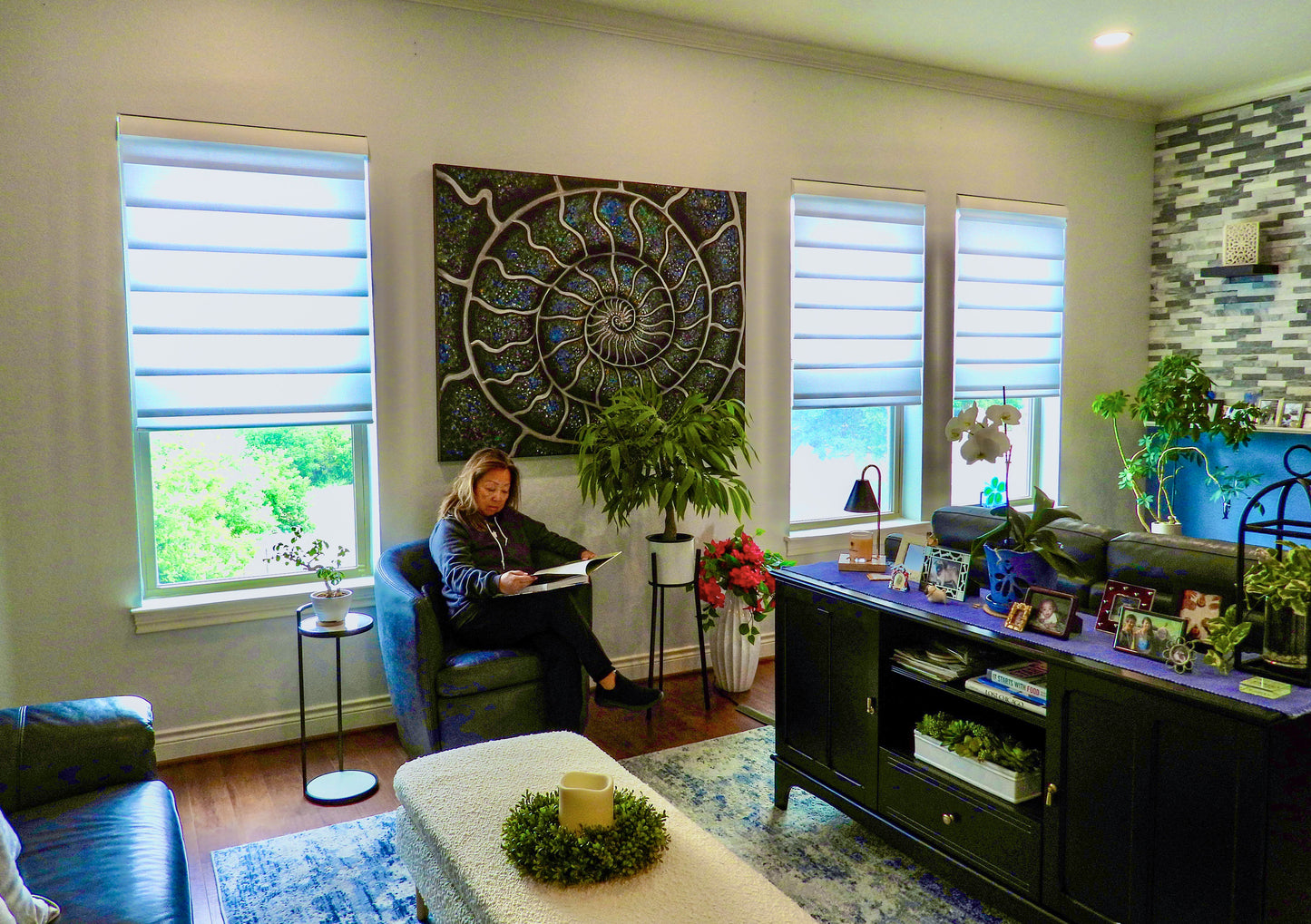 Living room with a woman sitting ina chair, surrounded by decorative elements. Behind her is a large abstract painting called Silver Blue Ammonite by Doug LaRue