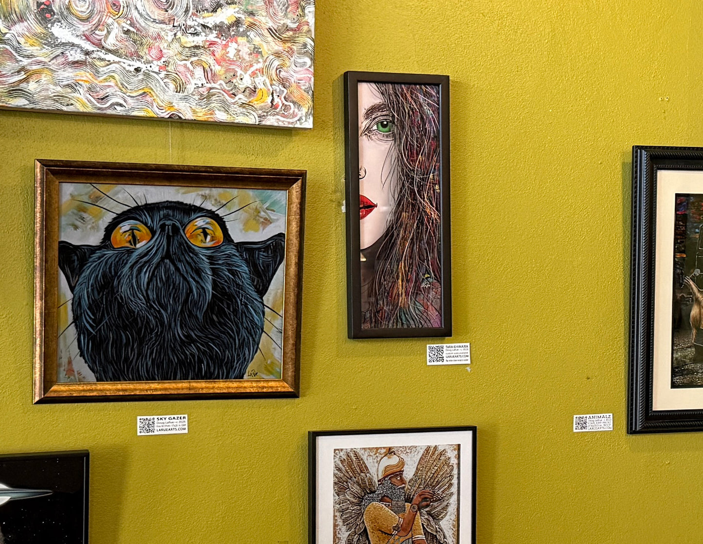 Artistic wall display with various framed artworks on a yellow wall. TAra's eye 19x9 art print in a black wood frame.