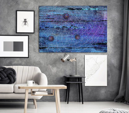 A large rectangular abstract wall art print featuring swirling blue, purple, and black tones with thin circular line patterns, displayed in a styled living room above a sofa and side table. Blue Trinity by Doug LaRue